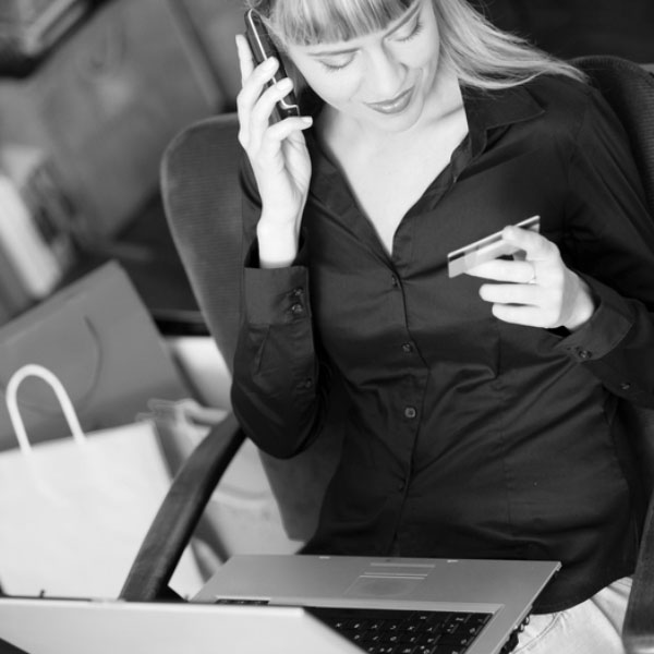 Black and white image of a woman talking on a phone and looking at her credit card with a laptop on her lap.