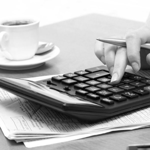 Black and white image of a a person's hand using a calculator that is sitting on papers with cup of tea in the background.