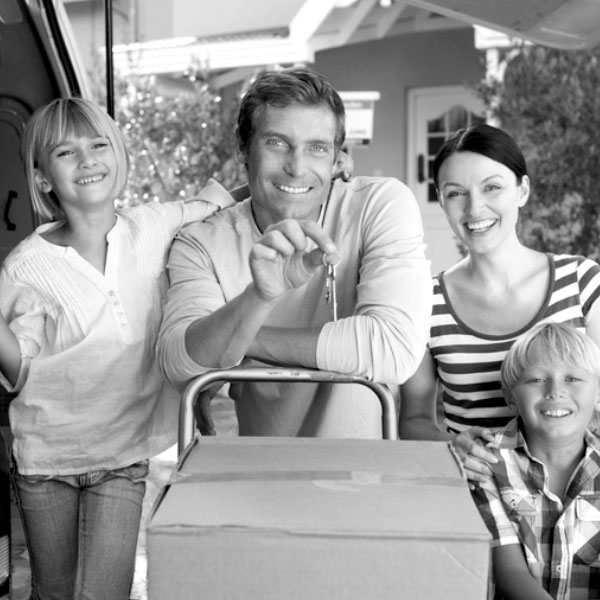 Black and white image of a family standing in front of a home. Dad is holding the key and leaning on a cart with a moving box on top.