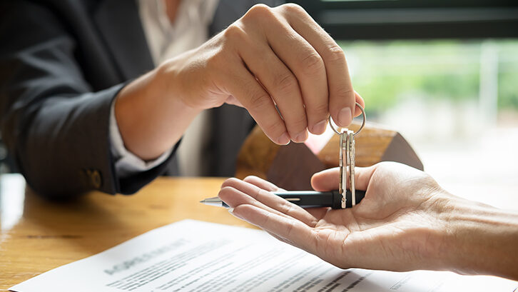 This photo represents buying a home or property in Bucks County, Pennsylvania. In the upper left corner, there is someone in a black suit jacket and white button down shirt. You can't see the person's head. They are reaching toward the center of the photo with a pair of silver keys on a small keyring in their hand. Another hand is held out from the bottom right corner, flipped over like they are going to catch the keys. On the table below is a blurred piece of paper.