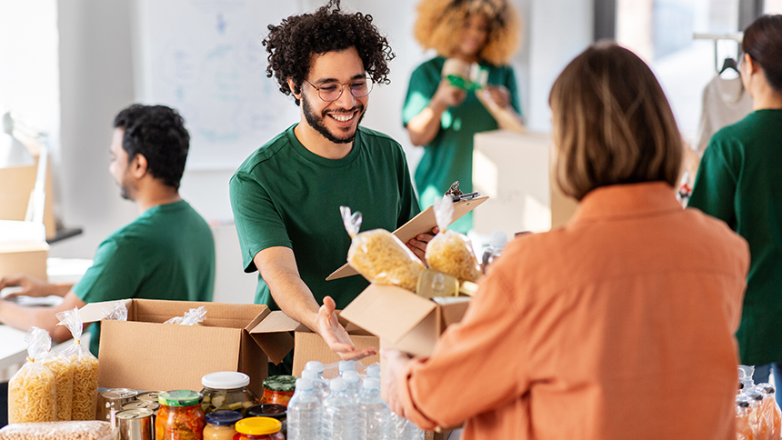 A group of five people are pictured at a nonprofit event. There are four volunteers are in green shirts. The image focuses on a man in his late 20s or early 30s reaching for a box of food from a woman in an orange shirt whose back is to the camera. There is a table they are both reaching over with pantry food laid across it. This image is to represent the corresponding blog, "Unlocking Opportunities: Nonprofit Low-Cost Financing Explained."
