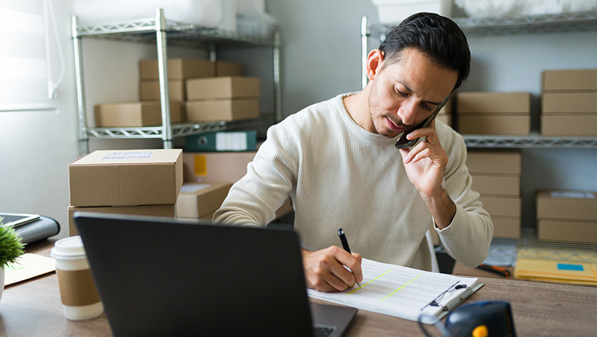 This photo represents the business loan program process through the Small Business Administration. The business owner is a man in his 30s and he is sitting at a table facing his laptop. He has a pen in his left hand and he is making notes on a piece of paper whil on the phone. There are boxes in the background behind him.