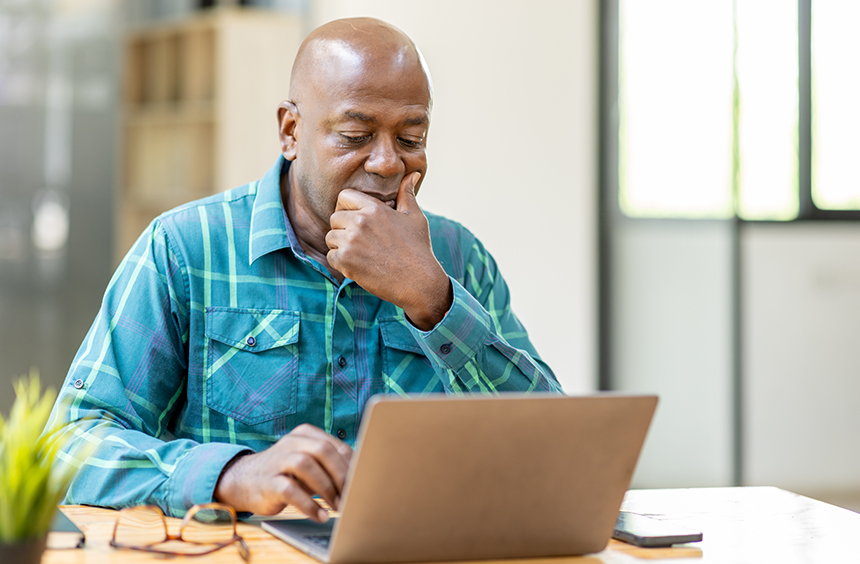 A photo to go with the blog, "The Benefits of Outsourcing Finances and Payroll." The image shows a manin a blue and green plaid button up shirt. He hold his left hand to his mouth in a pondering way, and his right hand hovers over the laptop keyboard he is looking at. 