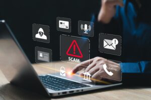 person sitting at a table with one hand on a laptop. identity theft/scam icons hovering above the laptop representing the importance of financial safety.